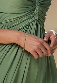 Vergulde armband met een gladde, gebogen vorm en een klein bedeltje, gedragen aan een hand die een groene, geplooide jurk van zachte stof vasthoudt.