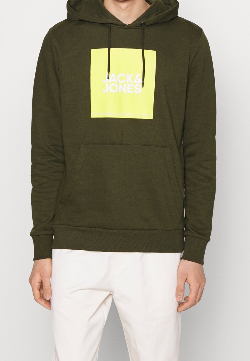 Sudadera de color verde oliva con un gran gráfico en forma de cuadrado amarillo que presenta "JACK & JONES" en blanco y "ESTABLECIDO 1990" debajo. Incluye un bolsillo frontal.