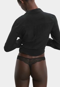 Zwarte kanten string met complexe bloempatronen, gecombineerd met een aansluitend zwart shirt dat een zachte textuur heeft en een licht cropped ontwerp.