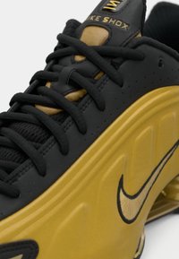 Μαύρο και χρυσό αθλητικό παπούτσι Nike Shox με μαύρες κορδόνες και ορατό λογότυπο Nike στη χρυσή σόλα.