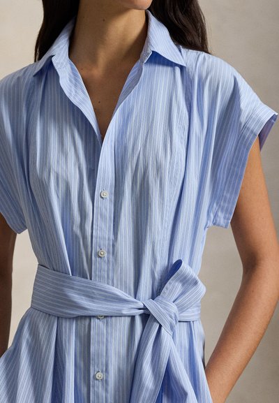 Φόρεμα πουκάμισο ανοιχτού γαλάζιου χρώματος με κάθετες λευκές ρίγες, κοντά μανίκια, γιακά και ζώνη που τονίζει τη μέση. Διαθέτει λεπτομέρειες με κουμπιά.