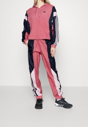 Kobieta ubrana w różny, granatowy i biały dres Adidas z bluzą z kapturem i pasującymi spodniami, w zestawie z czarnymi butami sportowymi Adidas.