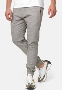 Homme portant un pantalon de jogging gris clair fuselé et des baskets blanches avec des accents vert olive et jaune, debout avec une jambe croisée.