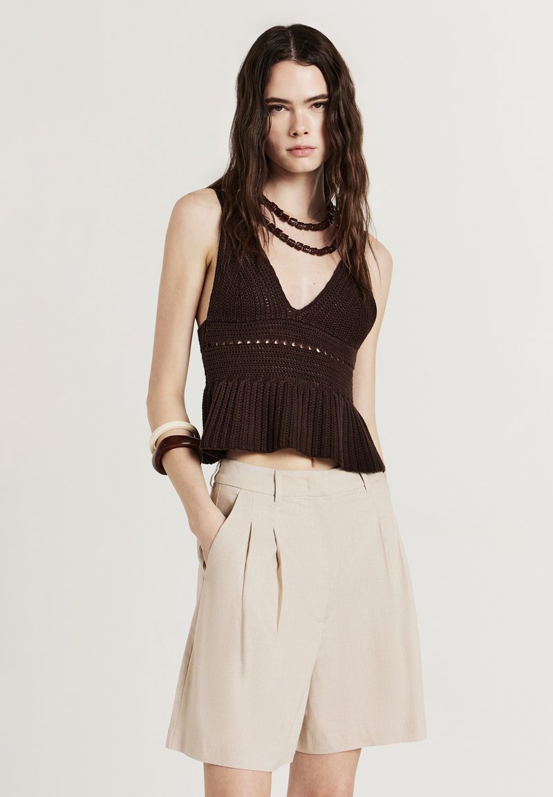 Donna che indossa un top smanicato in maglia marrone, pantaloni corti plissettati beige a vita alta, una collana a più strati e braccialetti, con le mani in tasca.