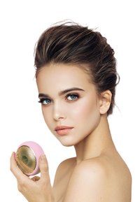 Rosa ansiktsrengöringsapparat med en guldtexturerad yta, hålls av en person. Slät, rundad form med en cirkulär design.