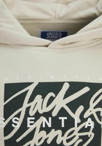Beige kapucnis pulóver, nagyméretű sötétzöld grafikával, fehér stilizált felirattal: "Jack & Jones" és "Essentials". Puha anyagú, laza fazon.