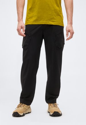 Άνδρας που φοράει μαύρο παντελόνι cargo με φερμουάρ στις πλευρικές τσέπες και μπεζ πεζοπορικά παπούτσια, στέκεται μπροστά σε λευκό φόντο.