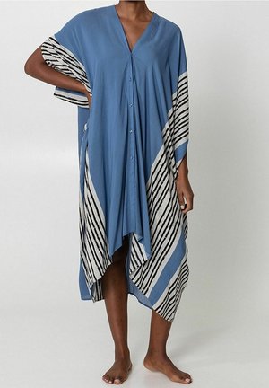 Kobieta ubrana w luźną, niebieską sukienkę do kolan z czarno-białymi ukośnymi paskami, stojąca boso na jasnym tle.