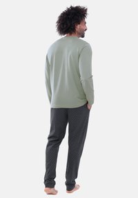 Hellgrünes Langarmshirt mit Rundhalsausschnitt, kombiniert mit dunklen Lounge-Hosen mit Diamantmuster. Barfuß.