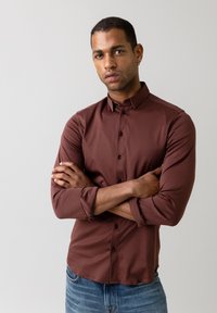 Cămașă maro, cu mânecă lungă, căptușită, cu un design ajustat. Textură netedă și manșete cu un singur buton. purtată cu blugi de culoare albastră deschisă.
