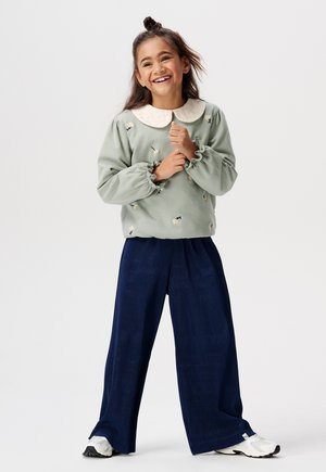 Lichtgroene sweatshirt met bloemborduring en een witte kraag, gecombineerd met marineblauwe ribbelachtige brede pijpen en witte sneakers.