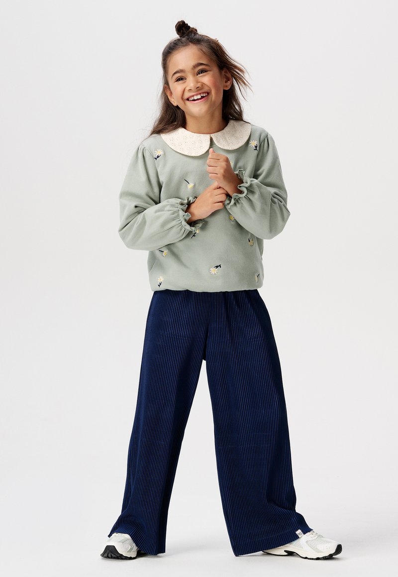 Lichtgroene sweatshirt met bloemborduring en een witte kraag, gecombineerd met marineblauwe ribbelachtige brede pijpen en witte sneakers.