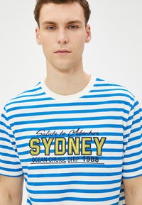 Niebieska i biała koszulka w paski z wyszytym napisem "SYDNEY OCEAN CRUISE SHIP 1988" w żółtych detalach. Okrągły dekolt, krótkie rękawy.