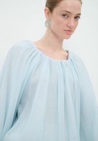 Himmelblaue, fließende Bluse mit weitem Ausschnitt und gerafften Details. Das Model trägt runde, verzierten Ohrringe. Weicher, strukturierter Stoff.