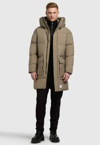 Giacca puffer beige con cappuccio, zip frontale e due tasche anteriori. Indossata sopra un top nero a collo alto e abbinata a pantaloni neri e stivali.