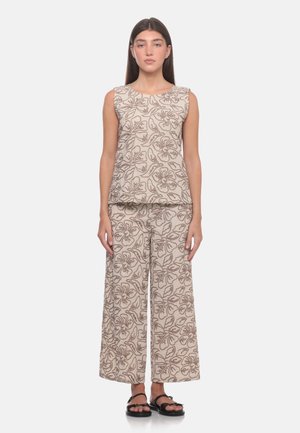 Donna che indossa un top beige senza maniche e pantaloni a gamba larga con motivi floreali marroni, in piedi a piedi nudi su sfondo bianco.