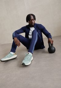 Completo di abbigliamento sportivo con giacca blu navy e pantaloni coordinati con accenti grigi, abbinati a scarpe azzurro chiaro e a un kettlebell nero.