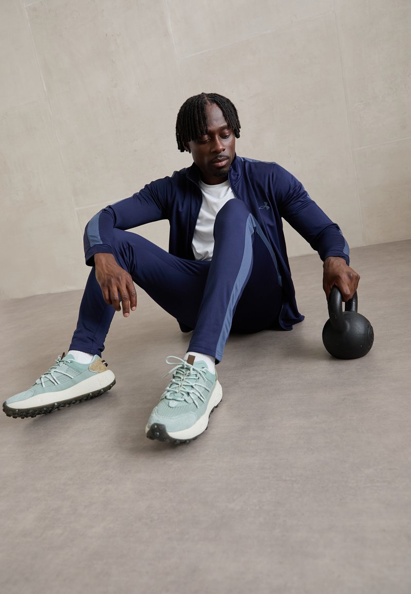 Ensemble de vêtements de sport comprenant une veste bleu marine et un pantalon assorti avec des accents gris, associé à des chaussures bleu clair et à un kettlebell noir.
