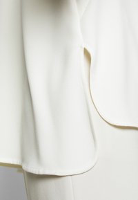 Blouse blanche en tissu léger et lisse ; présente un ourlet arrondi et de subtils fentes latérales pour un ajustement décontracté. Design épuré et minimal.