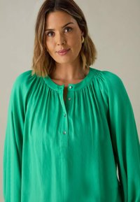 Blusa verde con scollo rotondo, design plissettato e chiusura con bottoni. Realizzata in tessuto leggero con una superficie liscia. Maniche lunghe.
