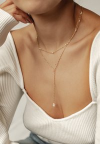 Goldfarbe Halskette mit kleinen Perlen, zarten Ketten und einem Y-förmigen Design mit einem Tropfenanhänger. Getragen über einem weißen gerippten Oberteil.