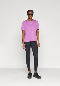 Μωβ αθλητική μπλούζα με κοντά μανίκια και μονόχρωμο σχέδιο. Συνδυασμένη με μαύρα κολάν και μαύρα αθλητικά παπούτσια. Το μοντέλο φοράει μαύρα γυαλιά ηλίου.