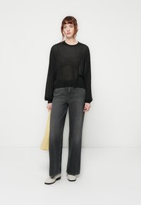 Haut noir transparent à manches longues associé à un jean gris taille haute et à jambes larges. Le modèle tient un sac jaune. Les chaussures sont blanches avec un talon massif.