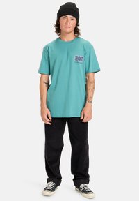 Mladý muž s tetováním, oblečený v tyrkysovém tričku s potiskem, černé čepici, černých kalhotách a černých teniskách s bílými tkaničkami, stojící na bílém pozadí.