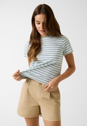 Vrouw met lang bruin haar, gekleed in een groen-wit gestreept T-shirt en beige korte broek, die de zoom van haar shirt rechtzet.