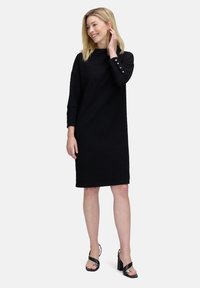 Schwarzes Strickkleid mit lockerer Passform, Dreiviertelärmeln mit dekorativen Knöpfen und knielangem Schnitt, kombiniert mit schwarzen Absatzsandalen.