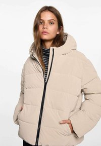 Beige puffjacka med huva, har en dragkedja fram och horisontellt quiltat mönster. Modellen bär en svartvit randig skjorta under.