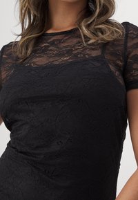 Kvinna som bär en svart spets topp med kort ärm med blommönster och ett enfärgat svart underlager, som visar mitt av torso och del av armen.
