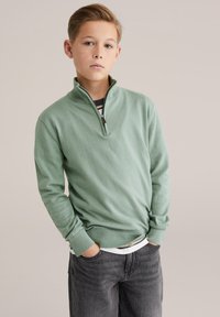 Pull vert clair à col zippé avec poignets et ourlet côtelés, porté par-dessus une chemise rayée, associé à un jean gris foncé. Texture lisse, design décontracté.