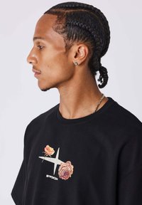 Camiseta de algodón negra con gráficos florales coloridos y un logo blanco; corte relajado con cuello redondo. Modelo mostrado de perfil.