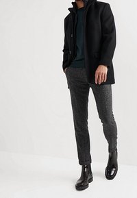 Černý kabát s vysokým límcem, přes tmavě zelený svetr, doplněný texturovanými šedými kalhotami a lesklými černými kotníkovými botami.