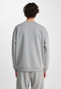 Uomo con i capelli ricci, che indossa un semplice felpa leggera grigia e pantaloni della tuta abbinati, in piedi con la schiena rivolta verso la telecamera.