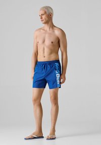 Homme torse nu aux cheveux blonds courts portant un short de bain bleu Red Bull et des tongs blanches, debout devant un fond gris uni.