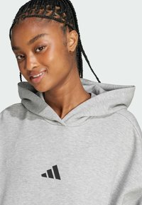 Sivi dukserica s velikim crnim Adidas logotipom na prsima, s kapuljačom na vezanje i mekanom teksturom. Glatka obrada i opušteni kroj.