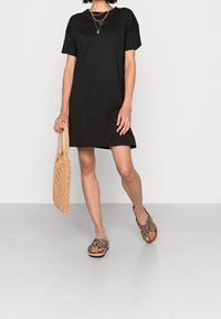 Svart t-shirtklänning med korta ärmar, tillsammans med sandaler med djurmönster och en vävd beige väska, på en neutral golvbakgrund.