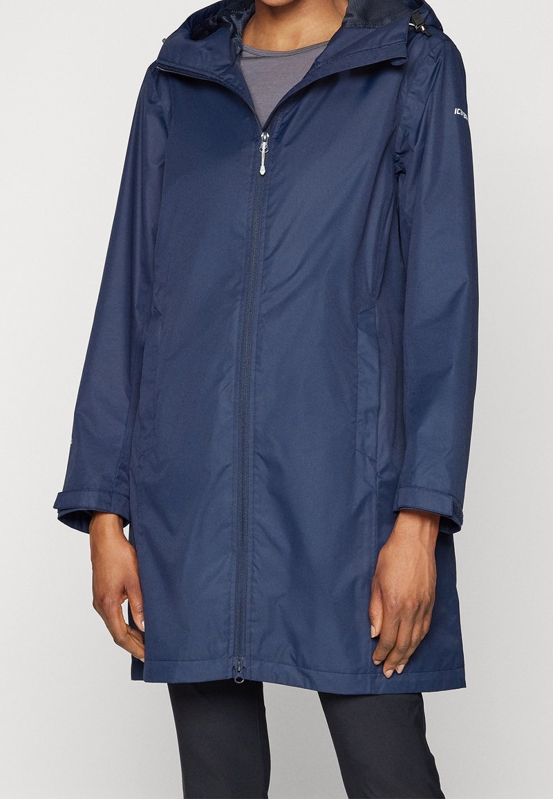 Personne portant un imperméable à capuche bleu marine fermé par une fermeture éclair, avec deux poches latérales et des poignets réglables, visible du cou jusqu'au milieu des cuisses.