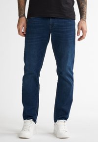 Jeans de mezclilla azul oscuro con un corte recto, con un ligero desgastado y detalles de costura, combinados con zapatillas blancas.