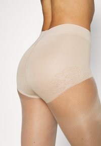Beige sømløse strømpebukser med et tekstureret blondemønster ved benåbningen. Letvægts, transparent materiale med en glat overflade.