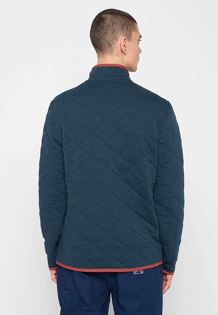 Giacca trapuntata navy con colletto alto, rifiniture rosse a contrasto e polsini abbinati. Tessuto morbido, leggero e dettagli a motivo a diamante.