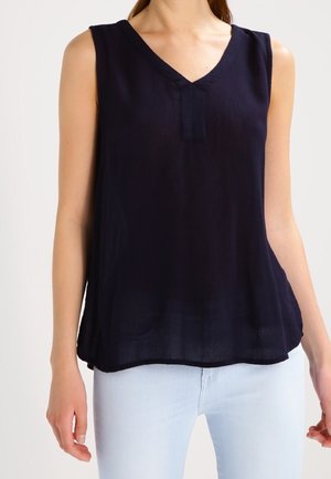 Kvinde iført sort, ærmeløs V-halsbluse kombineret med lyseblå jeans foran en ensfarvet baggrund.