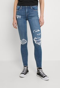 Jeans de mezclilla azul ajustados con múltiples zonas desgastadas y cortes deshilachados en las rodillas, combinados con zapatillas negras de caña alta.