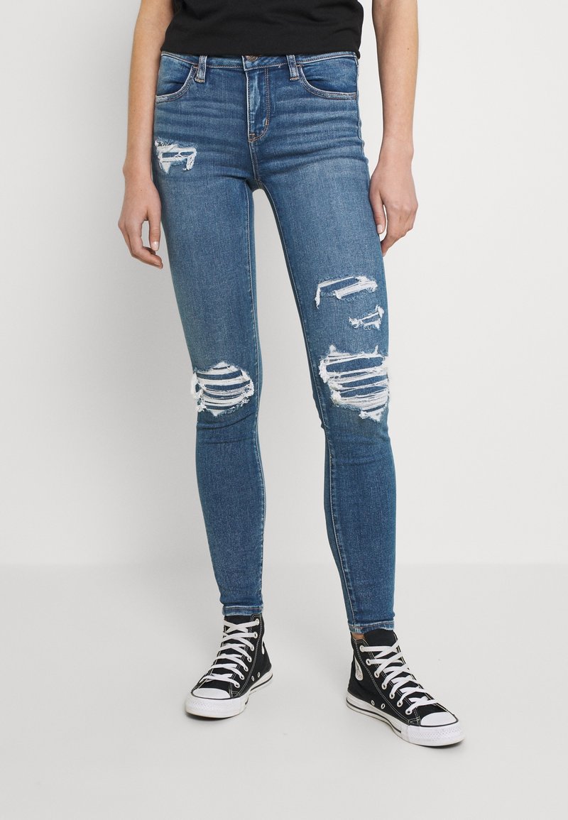 Jeans de mezclilla azul ajustados con múltiples zonas desgastadas y cortes deshilachados en las rodillas, combinados con zapatillas negras de caña alta.