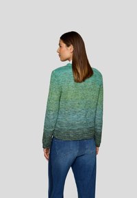 Frau mit glattem braunem Haar, die einen grün verlaufenden Strickpullover und blaue Jeans trägt, mit dem Rücken zu einem schlichten grauen Hintergrund gewandt.