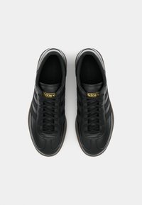 Sneaker in pelle nera con tre strisce scure, lacci piatti, accenti del logo dorati, punta rotonda e suola in gomma marrone a contrasto.