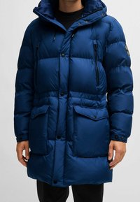 Blue puffer coat with a hood, featuring quilted sections, large front pockets, zippered chest pockets, and black button closures.