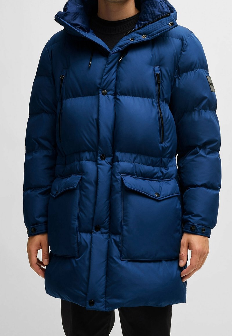 Blue puffer coat with a hood, featuring quilted sections, large front pockets, zippered chest pockets, and black button closures.
